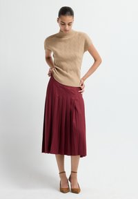 Top de punto beige con cuello alto y mangas cortas, combinado con una falda midi plisada en color burdeos. La modelo lleva zapatos de tacón marrones.