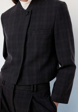 Gros plan d'une personne portant un blazer à carreaux foncés et un pantalon assorti avec un col montant et une fermeture à un bouton.