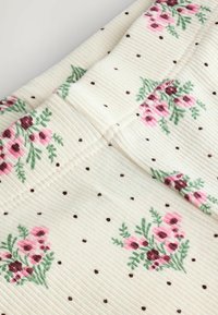 Weißer Stoff mit gerippter Textur, der verstreute braune Punkte sowie florale Muster in Rosa- und Grüntönen zeigt. Hervorstechende Nahtdetails sind sichtbar.