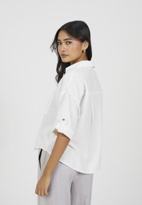 Kobieta z długimi ciemnymi włosami, nosząca złote kolczyki w kształcie kół, luźną białą koszulę zapinaną na guziki z podwiniętymi rękawami i jasne beżowe spodnie, stojąca bokiem.