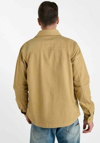 Veste beige avec un col et des poignets à boutons, présentant une empiècement arrière plat et un ourlet légèrement arrondi, associée à un jean bleu clair.
