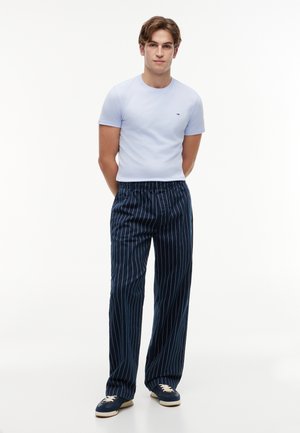 Jonge man staat met handen achter de rug, draagt een lichtblauw T-shirt, marineblauwe krijtstreeppanty en marineblauwe sneakers met witte veters.