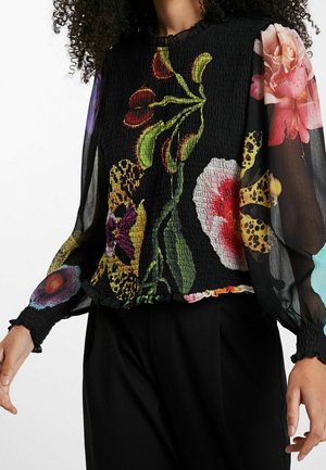 Mujer con blusa negra con grandes estampados florales coloridos y mangas abullonadas transparentes, combinada con pantalones negros.