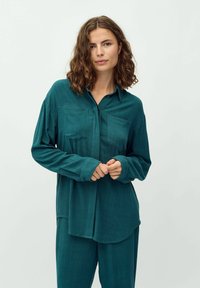 Camicia teal con bottoni, maniche lunghe, due tasche sul petto e un fit rilassato. Il tessuto ha un aspetto morbido e testurizzato.