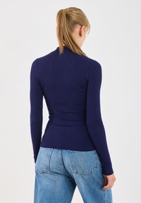 Kvinna med blont hår i en hästsvans som bär en åtsittande marinblå ribbad tröja och lösa blå jeans, vänd bort från en enkel vit bakgrund.