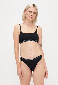 Femme aux cheveux blonds portant un bralette noir Hugo et un sous-vêtement noir Boss, se tenant devant un fond blanc uni.