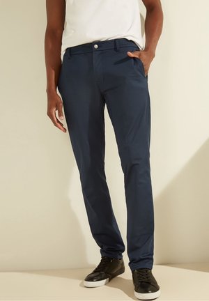 Man draagt marineblauwe slim-fit broek en zwarte sneakers met witte zolen, staand tegen een licht beige achtergrond.