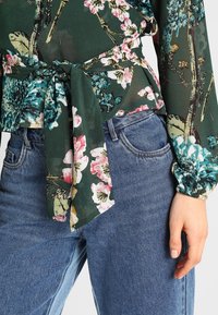 Långärmad blommig blus i mörkgrön, med rosa och blå mönster, och en knut detaljerat fram. Bar med högmidjade blå jeans.