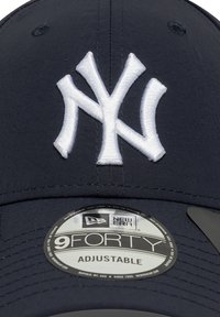 Jūras zila regulējama cepure ar baltu izšūtu "NY" logotipu. Gluda auduma, noapaļota forma un plakana mala ar New Era etiķeti.