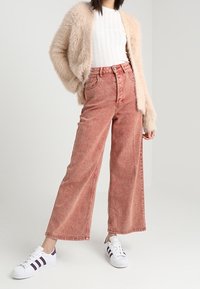 High-waisted, wide-leg jeans in faded coral denim, paired with a textured beige cardigan and white sneakers featuring black stripes.
