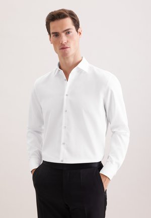 Mann trägt ein weißes Hemd mit Knöpfen und schwarze Hose, steht mit den Händen in den Taschen vor einem einfarbigen Hintergrund.