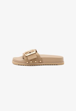 Sandalia slip-on beige con una correa texturizada, dos hebillas doradas y detalles de tachuelas. Suela plana de goma con un patrón de zigzag para mayor tracción.