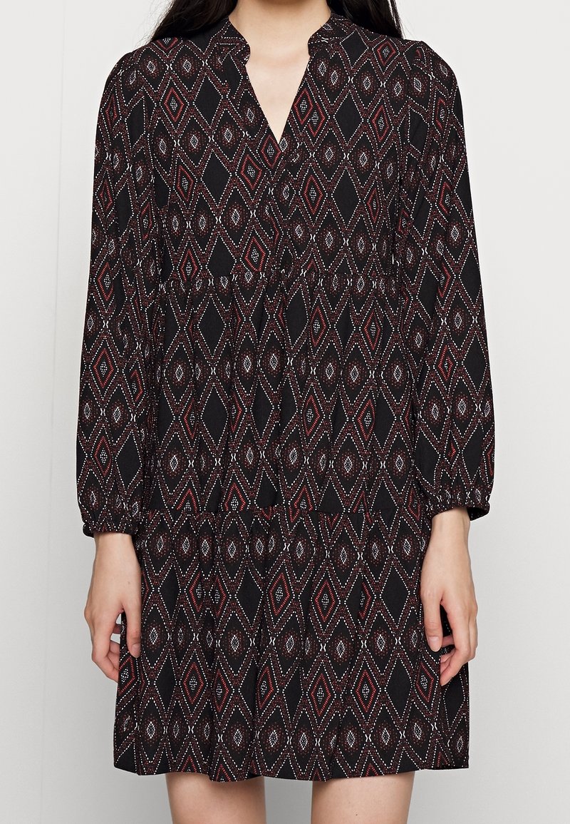 Schwarzes Kleid mit einem Diamantmuster in Rot und Weiß. Es hat lange Ärmel, einen V-Ausschnitt und ein design mit gestuften Röcken.