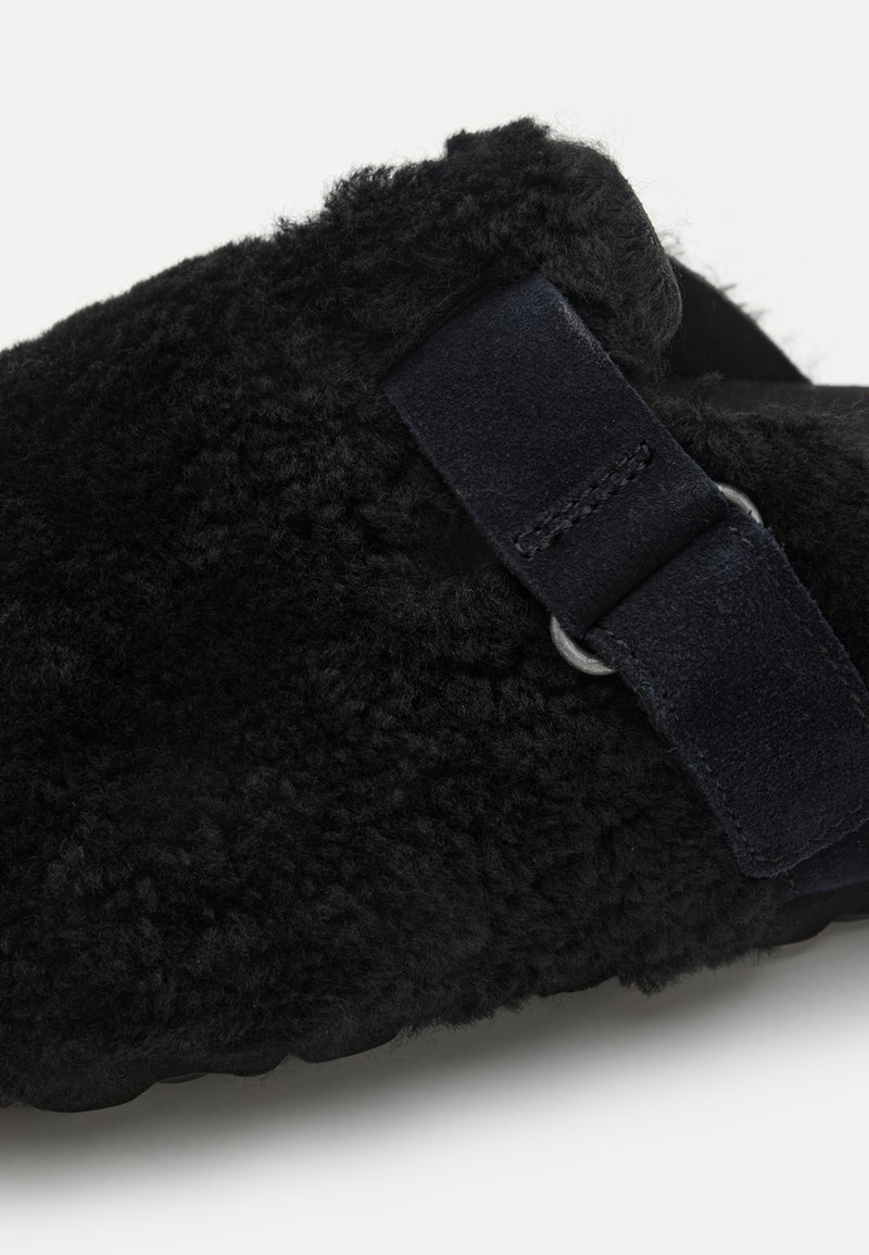 Schwarze Hausschuhe mit weichem, flauschigem Material, ausgestattet mit einem Wildleder-Riemen und silbernen Beschlägen. Das Design ist zum Hineinschlüpfen mit einer abgerundeten Form.