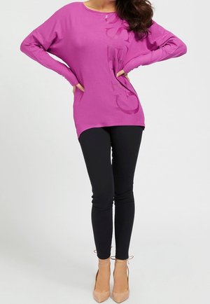 Vrouw die een paarse longsleeve met vage tekst draagt, zwarte leggings en beige hoge hakken, staand met haar handen in haar zij.