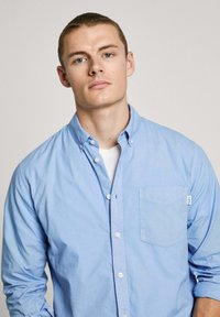 Ljust blå skjorta med knäppning, som har en bröstficka, knappned-krax, och långa ärmar med en slät yta.