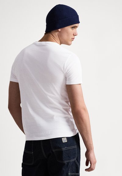 Bijela pamučna majica kratkih rukava klasičnog kroja, nošena s tamnoplavom pletenom kapom, uparena s plavim trapericama.