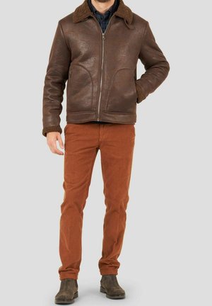 Uomo che indossa una giacca di shearling marrone, pantaloni color ruggine e stivali marrone scuro, in piedi con una mano in tasca.