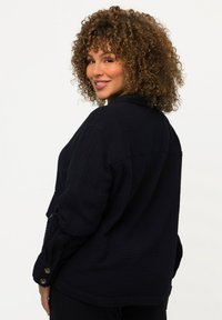 Zwarte textuur knoopshirts in een losse pasvorm, met een gebogen zoom en opgerolde mouwen, die een subtiel, gekreukt stofpatroon toont.