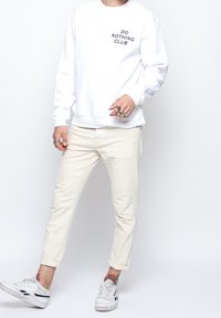 Bílá mikina s nápisem "DO NOTHING CLUB", béžové kalhoty a bílé tenisky. Neformální outfit s ohrnutými nohavicemi a viditelnými prsteny na rukou.