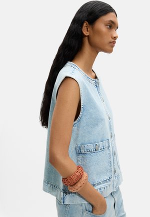 Kobieta o długich ciemnych włosach, ubrana w bezrękawnik z jasnoniebieskiego denimu i dżinsy, z dwoma strukturalnymi bransoletkami na nadgarstku.