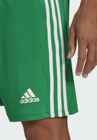 Pantalones cortos deportivos verdes con diseño de tres rayas blancas, que presentan un logo de Adidas bordado en blanco en la parte inferior izquierda. Tejido suave y texturizado.