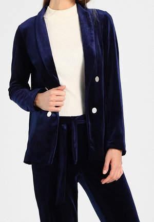 Vrouw draagt een marineblauw fluwelen pak met witte knopen over een crèmekleurige ribgebreide coltrui, met één hand houdt ze de revers van het jasje vast.