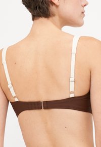 Barna melltartó fehér állítható pántokkal, sima textúrával és hátul kapcsos záródással. Egyszerű design, minta nélkül.