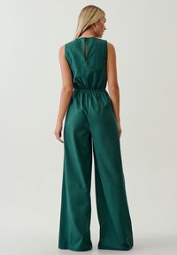 Tuta verde smeraldo con corpetto aderente e pantaloni a gamba larga, caratterizzata da un dettaglio a chiave sul retro e una vita elastica per il massimo comfort.