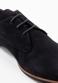 Tmavě námořnicky modrá semišová bota s kulatou špičkou, zdobená prošívaným vzorem a šedými tkaničkami. Podešev je kontrastní hnědé barvy.