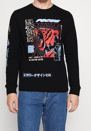 Maglietta nera a maniche lunghe con una grande grafica di una tigre ruggente in rosso e arancione, testo giapponese, codici a barre e accenti blu, indossata con jeans blu.