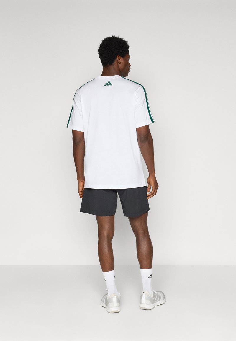 Witte sport t-shirt met groene strepen op de schouders, gecombineerd met donkere shorts. Het model draagt witte sokken en lichtgrijze sneakers.