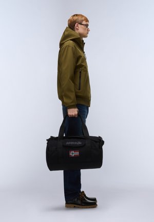 Joven con chaqueta oliva y gafas sosteniendo un bolso deportivo negro con la bandera de Noruega, de pie de lado contra un fondo liso.