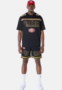 Jersey de malla negra con acentos dorados, que presenta el texto "49ERS San Francisco". Combinado con pantalones cortos negros a juego, que también muestran "49ERS". Gorra roja.
