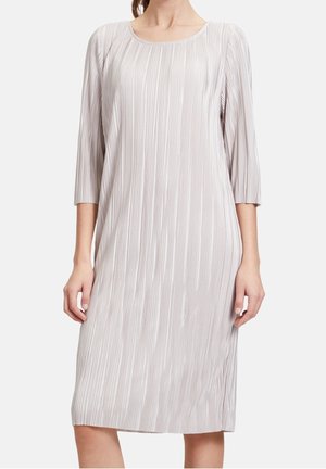 Frau trägt ein hellgraues, knielanges Kleid mit vertikalen Falten und dreiviertellangen Ärmeln vor schlichtem Hintergrund.