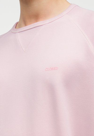 Gros plan sur une personne portant un sweat-shirt rose clair avec le texte brodé rose « CLOSED » sur la poitrine.