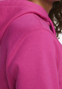 Fuchsiová mikina s kapucí z měkké, texturované látky. Má velkou kapuci a viditelné švy podél švů.