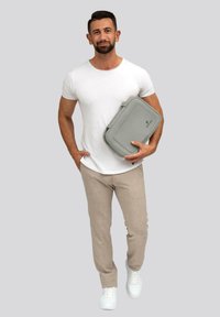 Borsa per laptop grigia con una superficie liscia, caratterizzata da bordi arrotondati e chiusura con zip. Il modello indossa una maglietta bianca e pantaloni beige chiaro.