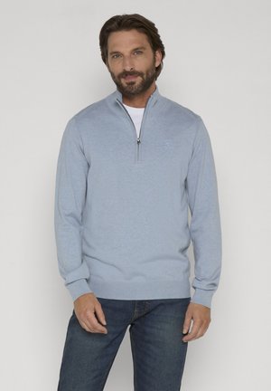 Mand iført lyseblå lynlås-sweater over hvid skjorte og blå jeans, stående mod en ensfarvet lys baggrund.