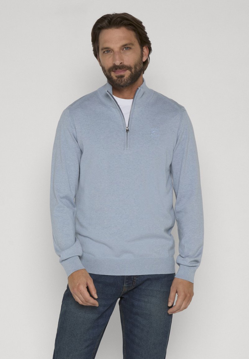 Homme portant un pull zippé bleu clair par-dessus une chemise blanche et un jean bleu, debout devant un fond clair uni.