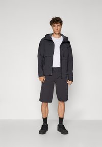 Veste noire ample avec une capuche et deux poches avant, associée à un short sombre. Les accessoires comprennent des baskets noires et des chaussettes courtes.
