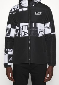 Svart och vit uppblåsbar jacka med geometrisk mönstring, logotypdetaljer, dragkedja fram och elastiska ärmslut. Texturerat tyg.