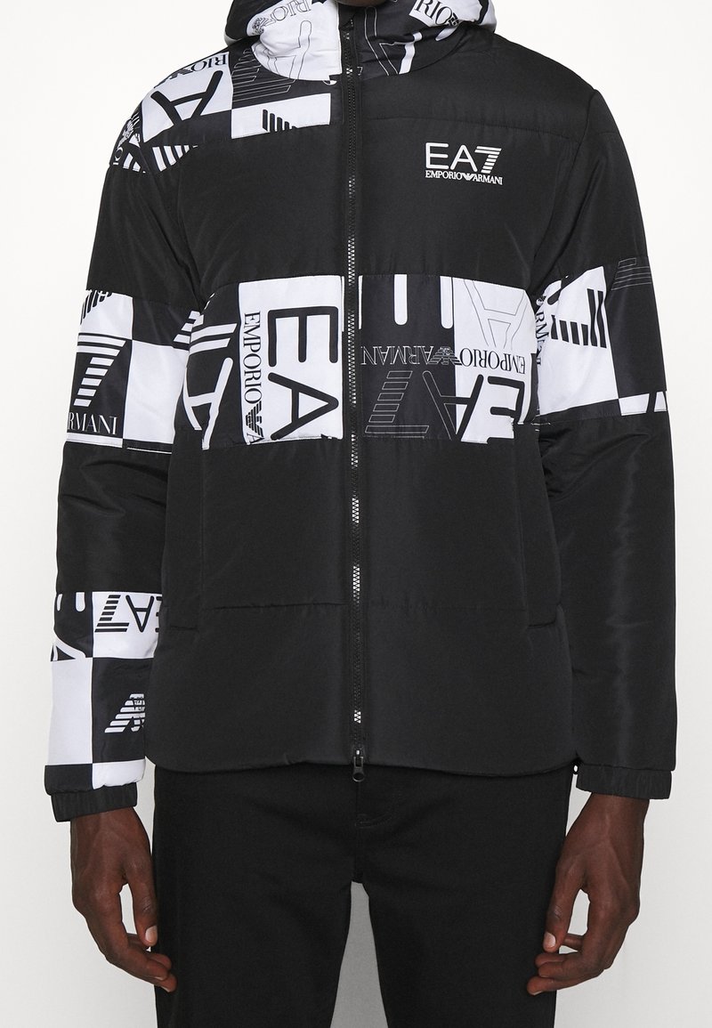 Svart och vit uppblåsbar jacka med geometrisk mönstring, logotypdetaljer, dragkedja fram och elastiska ärmslut. Texturerat tyg.