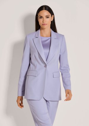 Vrouw met lang donker haar, gekleed in een licht lavendelkleurig op maat gemaakte blazer met bijpassende broek en een satijnen top, staand tegen een effen achtergrond.