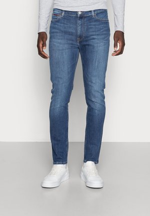 Mężczyzna w obcisłych niebieskich dżinsach i białych trampkach, stojący na jednolitym jasnym tle.