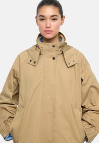 Beżowa kurtka z długim rękawem, z wysokim kołnierzem, z detalami guzikowymi, elastycznymi mankietami i bocznymi kieszeniami. Gładka tekstura, luźny krój.