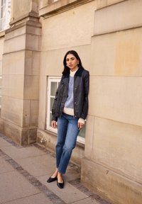 Das Model trägt eine schwarze leichte Jacke zu einem gestreiften Pullover in Blau und Creme, blaue gerade Jeans und schwarze Ballerinas. Es steht an einer Steinmauer.