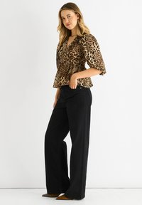 Punkte mit Leopardenmuster in einem taillierten Oberteil mit kurzen Ärmeln; kombiniert mit schwarzen, weiten Hosen und beige-farbenen, spitzen Schuhen. Heller Hintergrund, Model seitlich positioniert.