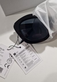 Mustad mustakallisilmalised prillid tumedate läätsedega. Kaasas sildid, mis näitavad hooldusjuhiseid ja CE sertifitseerimise üksikasju.