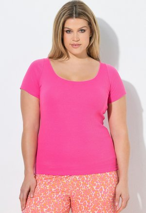 Donna con lunghi capelli biondi che indossa una maglietta rosa acceso a maniche corte e pantaloni rosa e arancioni con fantasia, in piedi contro un muro bianco.